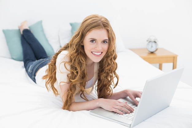 Sonriendo casual rubia usando la computadora portátil en la cama