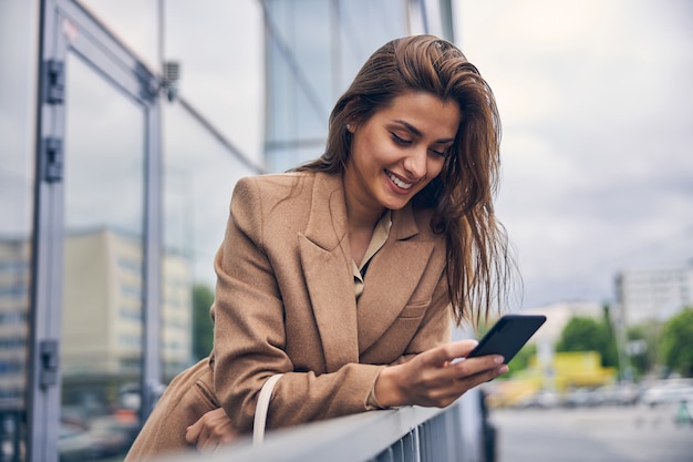 Sonriendo complacido elegante apuesto joven empresaria caucásica con un gadget apoyado en el riel