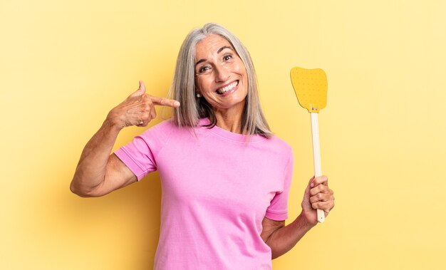 Sonriendo con confianza apuntando a su propia sonrisa amplia, actitud positiva, relajada y satisfecha y sosteniendo un matamoscas