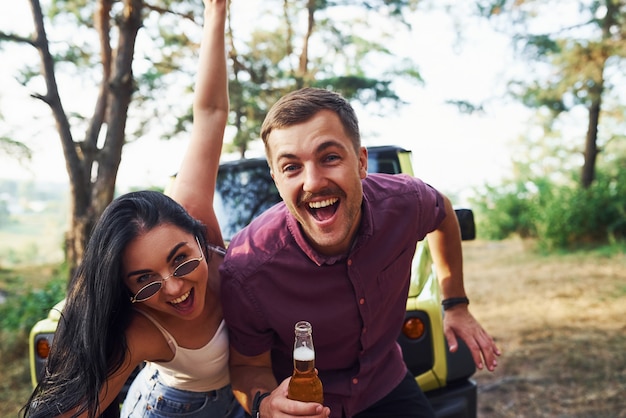 Sonriendo y divirtiéndose. Pareja de jóvenes con alcohol se divierten en el bosque. Jeep verde detrás.