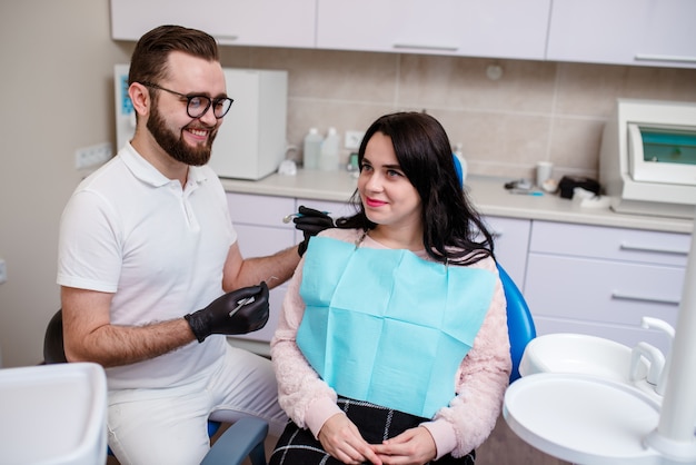 Sonriendo doctora y su asistente mostrando los rayos x de los dientes al paciente masculino en el hospital dental. Paciente masculino hermoso que se sienta en la silla smilling con los dientes blancos rectos y que mira la radiografía dental.