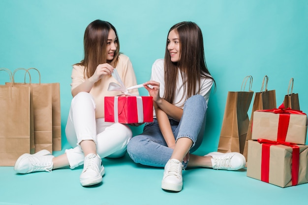 Sonriendo a dos niñas jóvenes sentados en el suelo bolsas de compras y regalos abiertos en la pared turquesa.