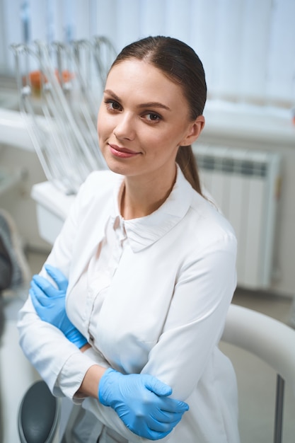 Sonriendo a ella dentista en uniforme médico y guantes estériles disfrutando de su día de trabajo en la oficina moderna