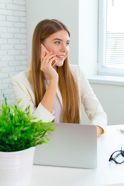 Sonriendo empresaria sentada en su lugar de trabajo en la oficina y hablando por teléfono