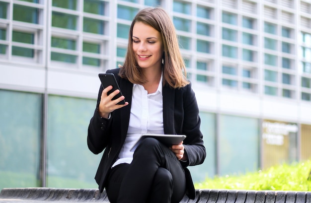 Sonriendo empresaria usando una tableta digital al aire libre sentado en un banco y susing también su teléfono inteligente