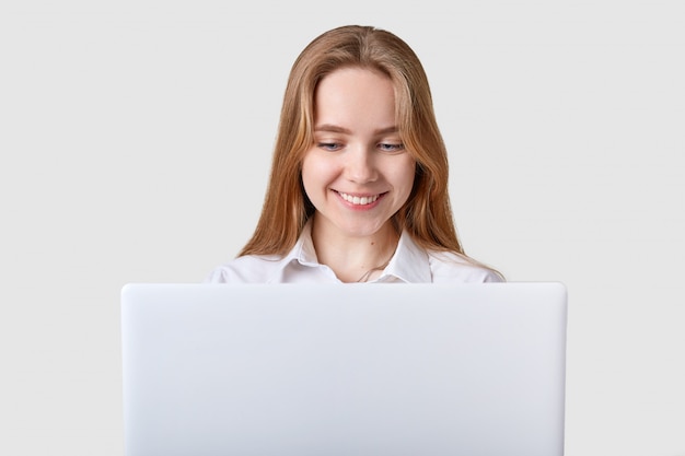 Sonriendo encantada niña de ojos azules se sienta a la mesa, trabajando en la computadora portátil
