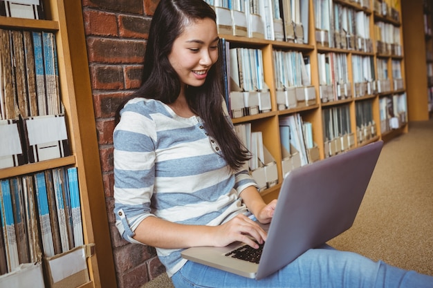 Sonriendo estudiante sentado en el piso contra la pared en la biblioteca usando laptop