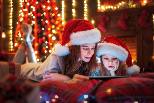 Sonriendo familia madre e hija en santas sombreros y pijamas viendo videos graciosos o eligiendo regalos en tableta digital