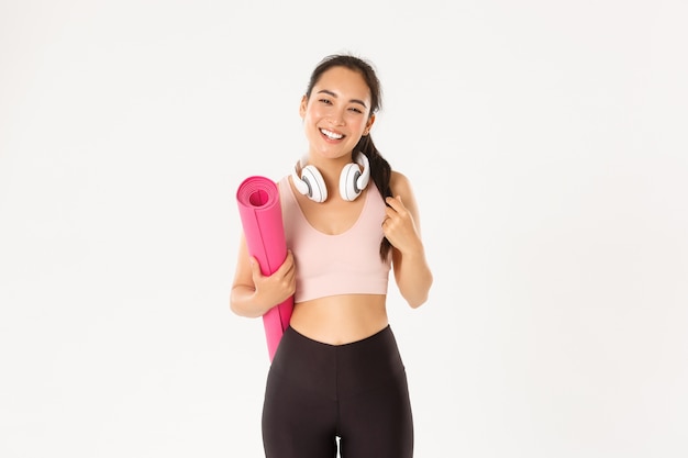 Sonriendo feliz chica asiática fitness en auriculares y ropa deportiva, llevar alfombra de goma para entrenamiento o yoga, riendo sin preocupaciones sobre fondo blanco.