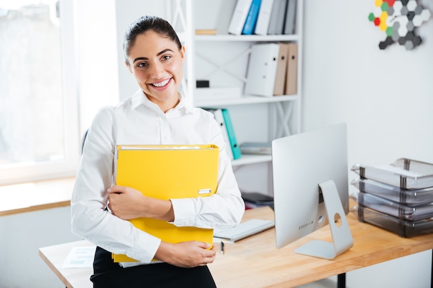 Sonriendo feliz empresaria sentada en la mesa con documentos en la oficina y mirando al frente