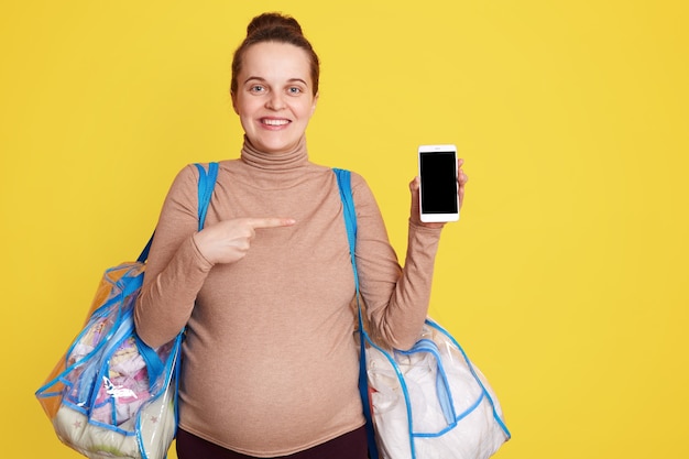 Sonriendo feliz futura madre sosteniendo bolsas con cosas para dar a luz, posando contra la pared amarilla con el teléfono en las manos y apuntando a la pantalla en blanco con el dedo índice.