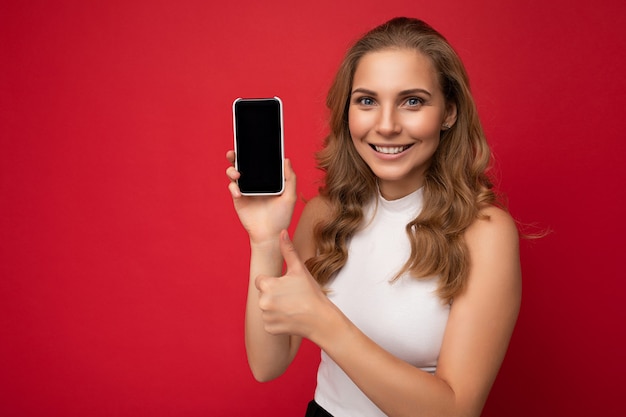 Sonriendo feliz joven y bella mujer rubia vistiendo camiseta blanca aislado en superficie roja