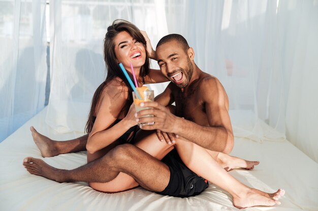 Sonriendo feliz pareja multirracial sentada en la cama y bebiendo en la playa