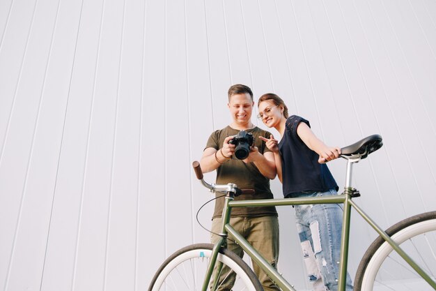Foto sonriendo fotógrafo y mujer feliz revisando fotos después de la sesión de fotos, de pie con bicicleta retro sobre pared blanca. al aire libre.