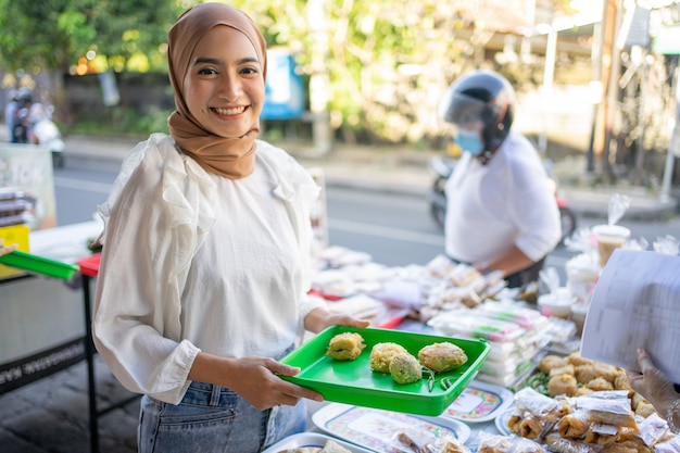 Sonriendo a una hermosa niña en un velo mantenga una bandeja de plástico vendiendo varios tipos de alimentos fritos