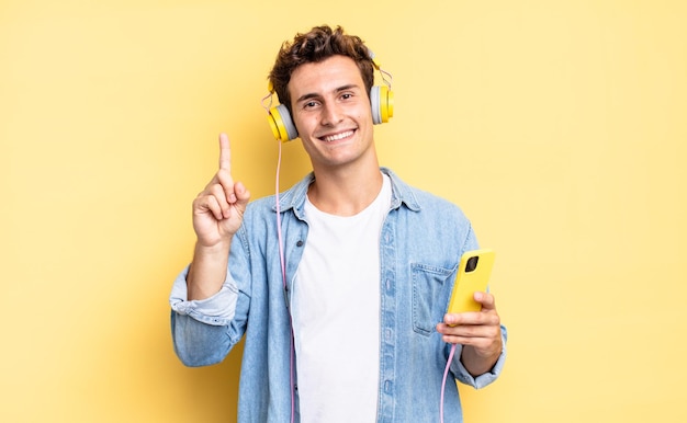 Sonriendo y luciendo amigable, mostrando el número uno o el primero con la mano hacia adelante, contando hacia atrás. concepto de auriculares y teléfono inteligente