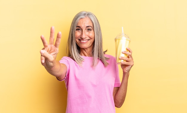 Sonriendo y luciendo amigable, mostrando el número tres o tercero con la mano hacia adelante, contando hacia atrás y sosteniendo un batido.