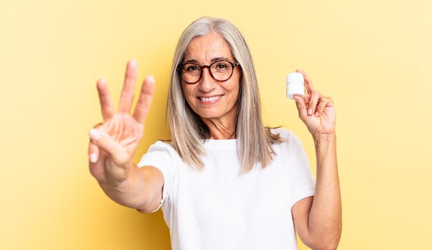 Sonriendo y luciendo amigable, mostrando el número tres o tercero con la mano hacia adelante, contando hacia atrás y sosteniendo un frasco de pastillas