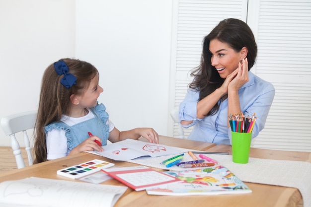 Foto sonriendo madre e hija preparándose para lecciones y dibuja en la mesa con lápices y pinturas