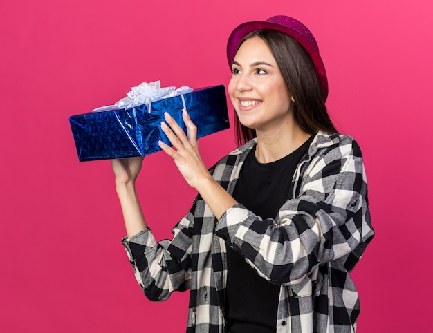 Sonriendo mirando al lado hermosa joven vistiendo gorro de fiesta con caja de regalo