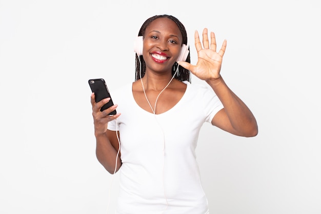 Sonriendo y mirando amistosamente, mostrando el número cinco con auriculares y un teléfono inteligente.