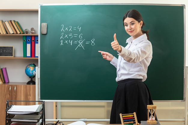 Sonriendo mirando el lado mostrando el pulgar hacia arriba joven maestra de pie detrás de la mesa con puntos de herramientas escolares con la mano en la pizarra en el aula