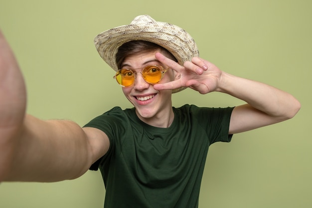 Sonriendo mostrando gesto de paz chico guapo joven con sombrero con gafas aislado en la pared verde oliva