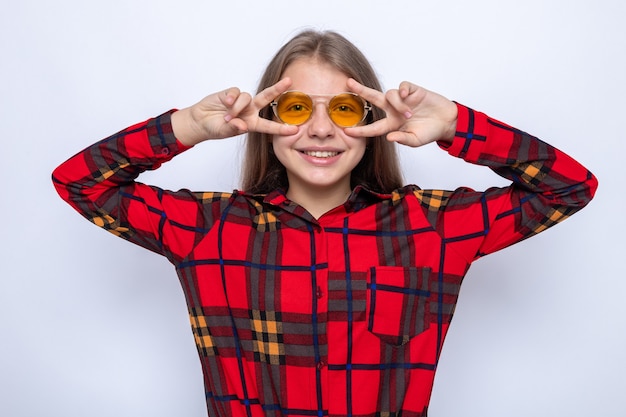 Sonriendo mostrando gesto de paz hermosa niña con camisa roja y gafas aisladas en la pared blanca