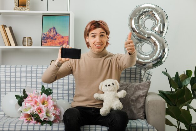 Sonriendo mostrando el pulgar hacia arriba chico guapo en el día de la mujer feliz con oso de peluche con teléfono sentado en el sofá en la sala de estar