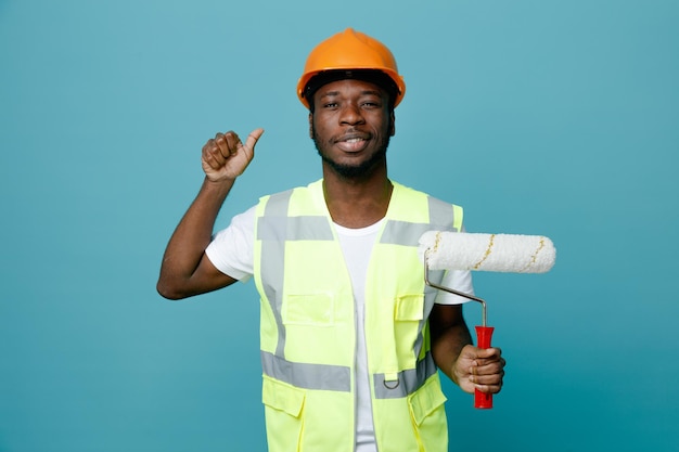 Sonriendo mostrando los pulgares hacia arriba joven constructor afroamericano en uniforme sosteniendo cepillo de rodillos aislado sobre fondo azul.