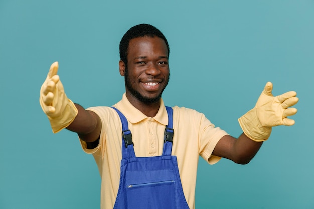 Sonriendo mostrando tamaño joven limpiador afroamericano macho en uniforme con guantes aislado sobre fondo azul.