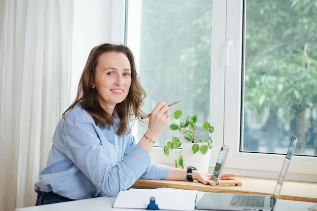 Sonriendo a la mujer de negocios de la cámara con camisa azul sentada detrás de un escritorio con una computadora portátil