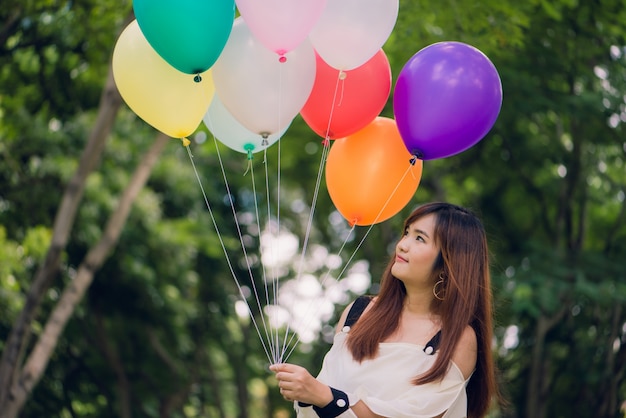 Sonriendo a las mujeres asiáticas hermosas jovenes con el pelo marrón largo en el parque. Con los globos de aire de color arco iris en sus manos. Una energía solar y positiva de la naturaleza.