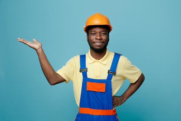 Sonriendo poniendo las manos en los puntos de las caderas al lado joven constructor afroamericano en uniforme aislado sobre fondo azul.