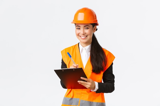 Sonriendo satisfecha ingeniera de construcción asiática líder de inspección en la empresa, con casco de seguridad y chaqueta reflectante, anotando notas y mirando el resultado satisfecho, fondo blanco.