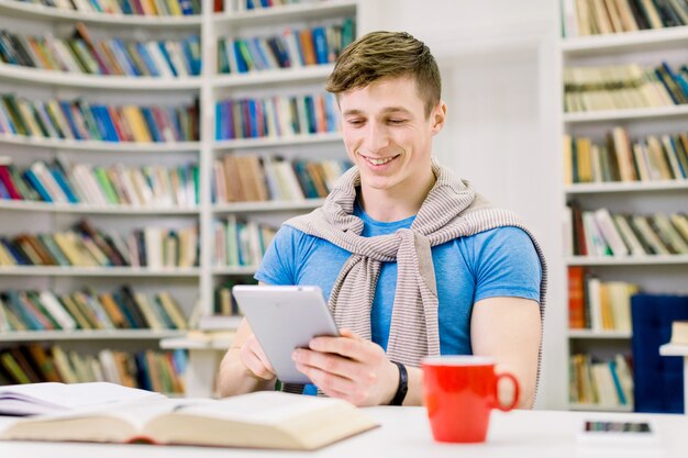 Sonriendo satisfecho guapo joven estudiante masculino sentado en la biblioteca y buscando información en i-pad durante la preparación para el examen