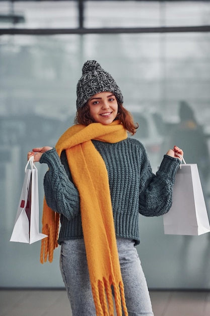 Foto sonriendo y sintiéndose feliz. hermosa chica alegre con bufanda amarilla y ropa de abrigo parada en el interior con bolsas de compras en las manos.