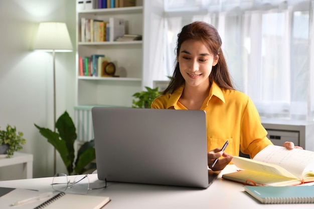 Sonriendo y sintiéndose positivo. feliz joven mujer asiática independiente trabajando en la computadora en casa. Atractiva mujer de negocios que estudia en línea, usa software de computadora portátil, navega por la web o compra en