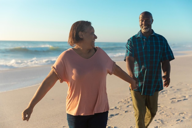Sonriente anciana afroamericana sosteniendo la mano del hombre caminando en la playa