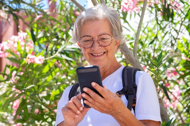 Sonriente anciana con anteojos sosteniendo el teléfono móvil mientras está de pie bajo una planta florecida día soleado de verano