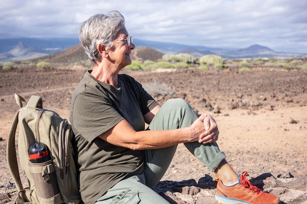 Sonriente anciana en caminata al aire libre sentada en un muro de piedra mirando hacia otro lado Mujer madura activa sentada cerca de su mochila disfrutando de la libertad y el día soleado