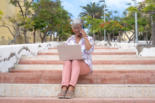 Sonriente anciana con camisa floreada sentada al aire libre en una escalera en la ciudad usando una laptop Caucásica atractiva mujer adicta a la tecnología y social