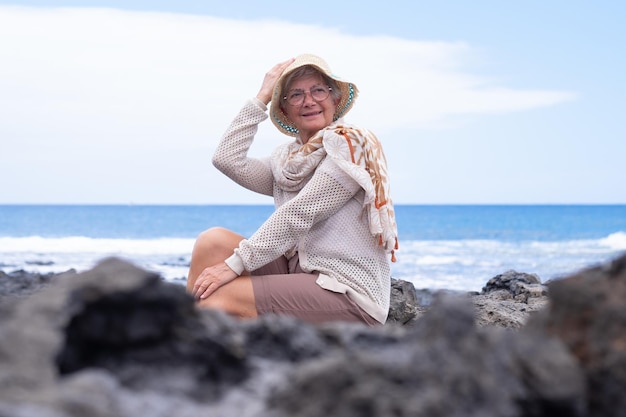 Sonriente anciana caucásica con sombrero sentado en la playa mirando hacia otro lado disfrutando de vacaciones o jubilación Horizonte sobre el espacio de copia de agua