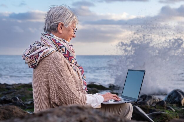 Sonriente anciana caucásica en trabajo remoto sentada en la playa usando una computadora portátil Anciana de cabello blanco disfrutando de vacaciones en el mar escribiendo en la computadora