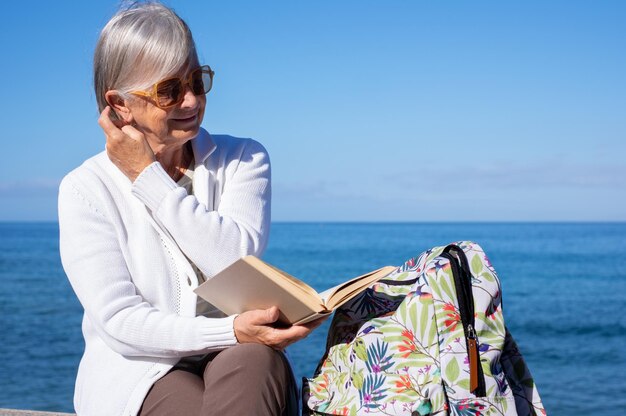 Sonriente anciana de pelo gris sentada relajada cerca de la playa disfrutando leyendo un libro