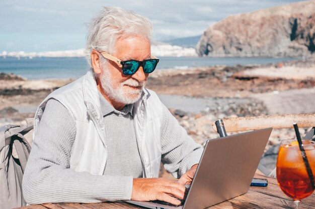 Sonriente anciano barbudo usando computadora portátil al aire libre en la playa en un día soleado