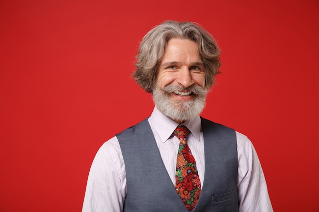 Sonriente anciano canoso bigote barbudo hombre en camisa clásica chaleco colorido corbata posando aislado sobre fondo rojo, retrato de estudio. Gente emociones sinceras concepto de estilo de vida. Simulacros de espacio de copia.