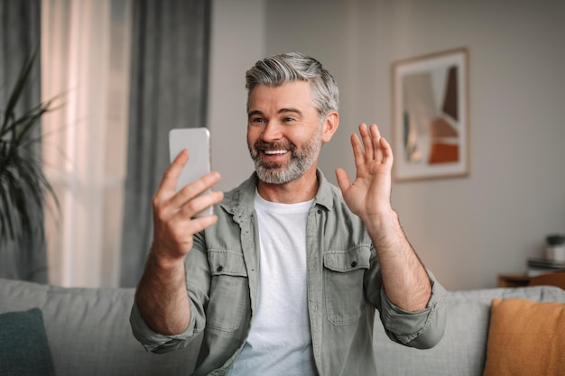 Sonriente anciano europeo con barba agitando su mano en la cámara web del teléfono inteligente en el interior de la sala de estar