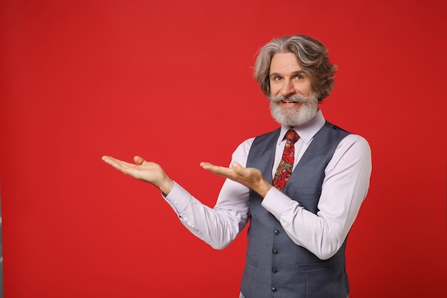 Sonriente anciano de pelo gris con bigote barbudo en chaleco de camisa clásico y corbata colorida aislado en un retrato de estudio de fondo rojo. Concepto de estilo de vida de las personas. Simulacros de espacio de copia. Apuntando las manos a un lado.