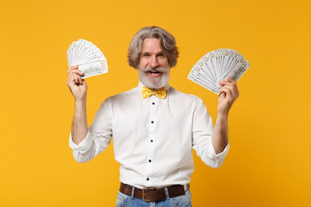 Sonriente anciano de pelo gris bigote barbudo hombre con camisa blanca pajarita aislado sobre fondo amarillo naranja. Concepto de estilo de vida de las personas. Simulacros de espacio de copia. Mantenga fan de dinero en efectivo en billetes de dólar.